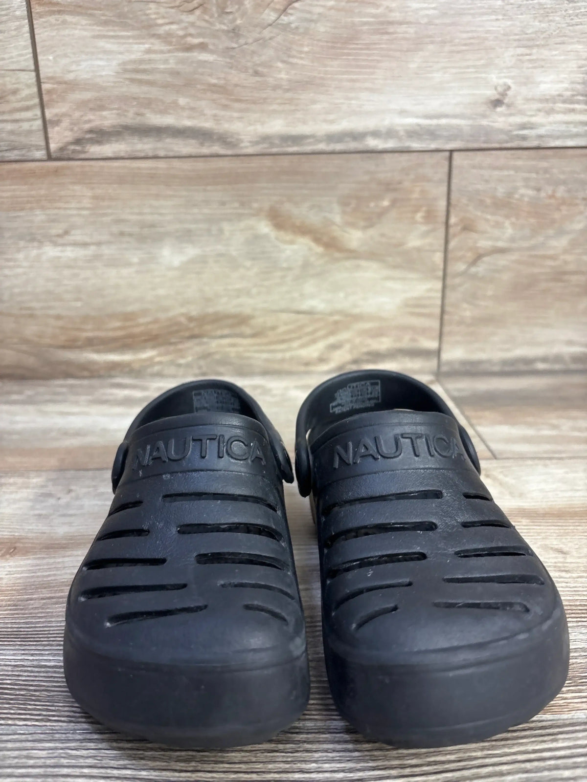 Front view of Nautica River Edge clogs in black showing ventilation slits and molded strap with Nautica logo on wood floor
