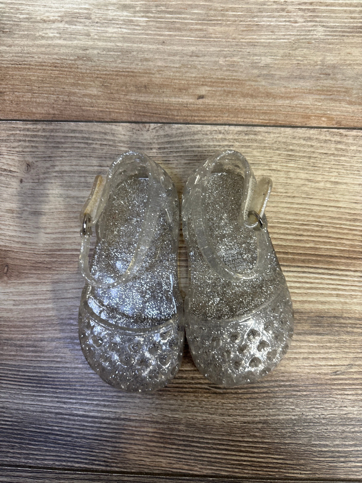 Top-down view of Old Navy toddler clear jelly sandals showcasing silver glitter finish and ankle straps on wood floor