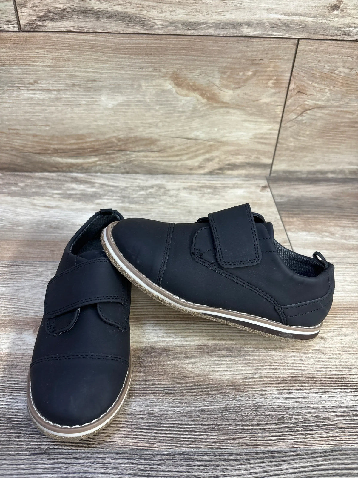 Angled front and side view of toddler black derby shoes with Velcro strap and stitched tan-and-white sole on wood background