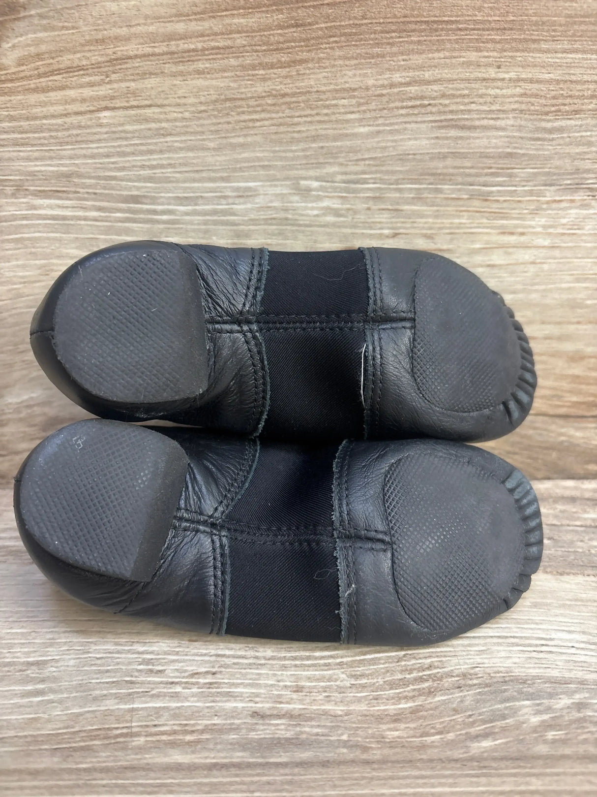 Bottom view of Capezio EJ2C girl’s black jazz slip-on shoes showing split rubber toe and heel soles on wood background