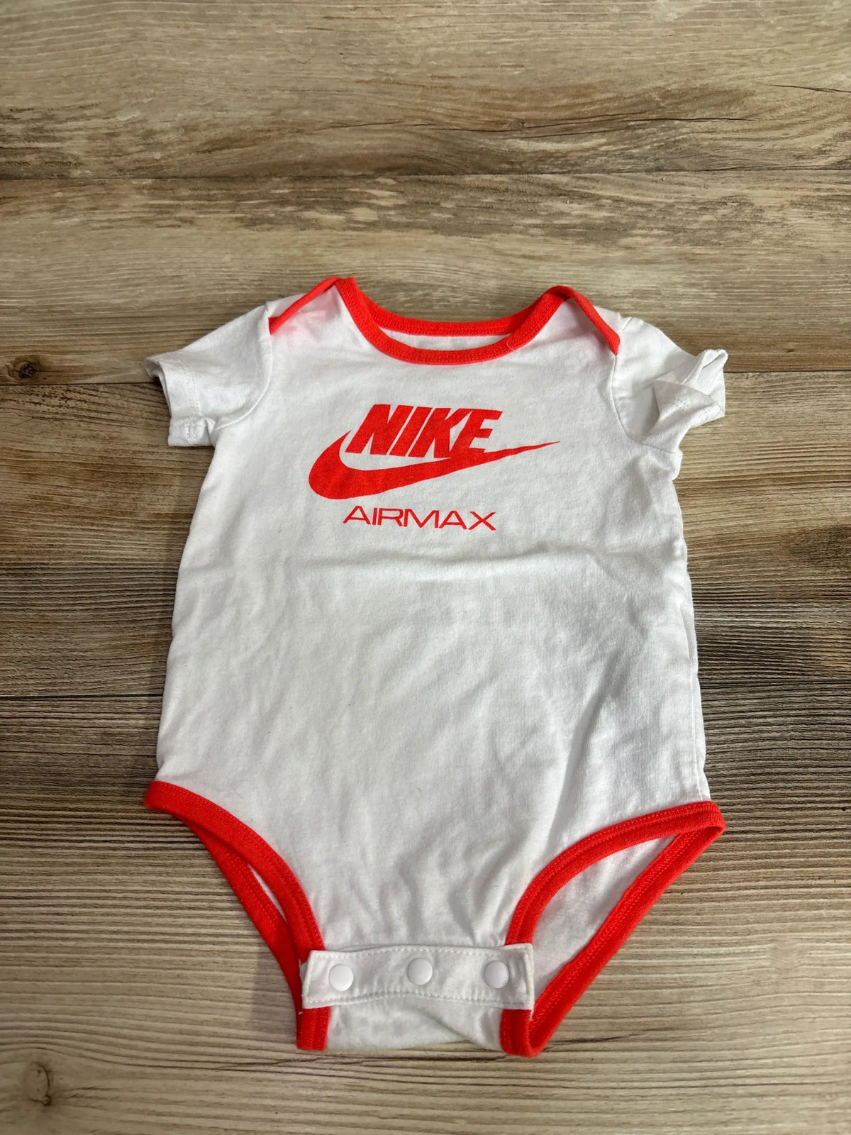 Front view of white Nike Airmax bodysuit, size 6 to 12 months, showcasing red trim, logo, and snap closures on wood grain