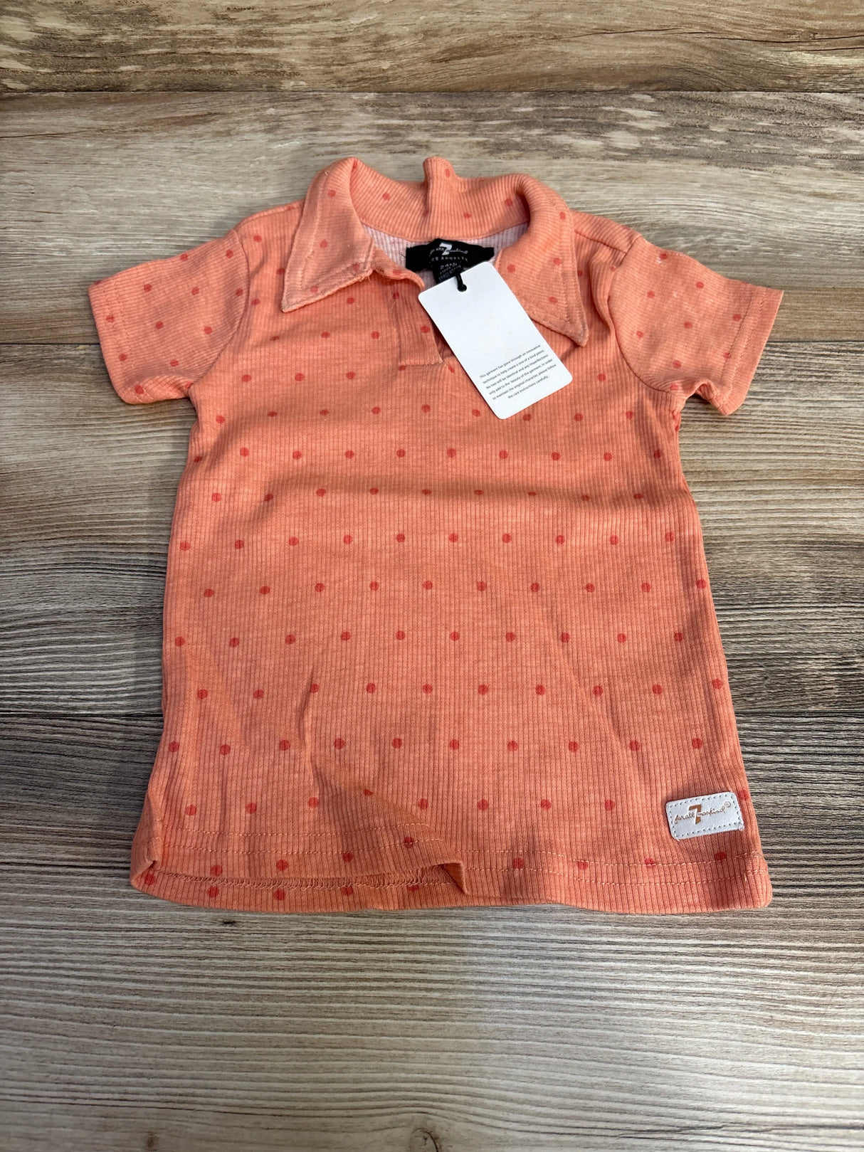 Flat lay of NWT 7 For All Mankind ribbed polo shirt in coral orange with red polka dot print and hangtag on wood plank floor