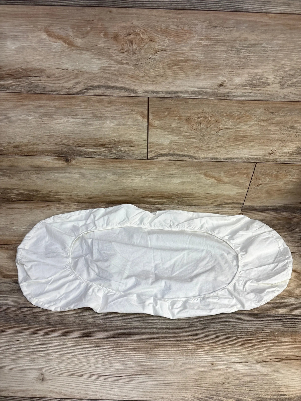 Flat lay of Happiest Baby SNOO Bassinet Sheet in white showcasing fitted elastic edges on wood plank floor