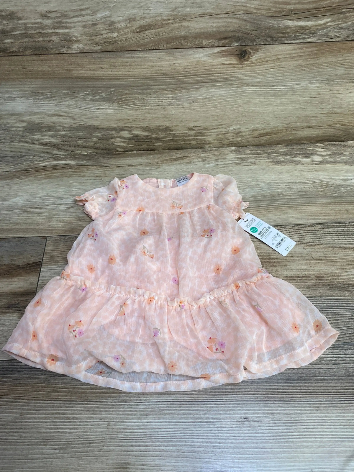 Front flat-lay of pale pink floral baby dress with ruffled hem and short puff sleeves on wooden floor background
