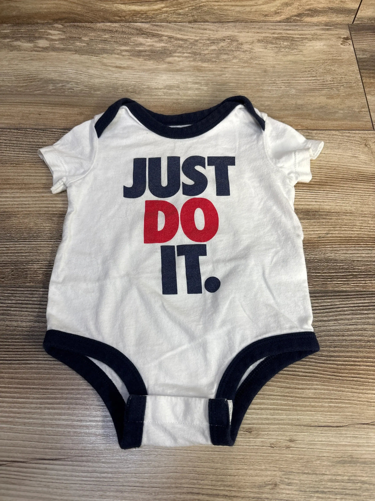 Top-down view of white baby bodysuit (6M) with navy trim and crotch snaps, featuring large JUST DO IT. text on wood background