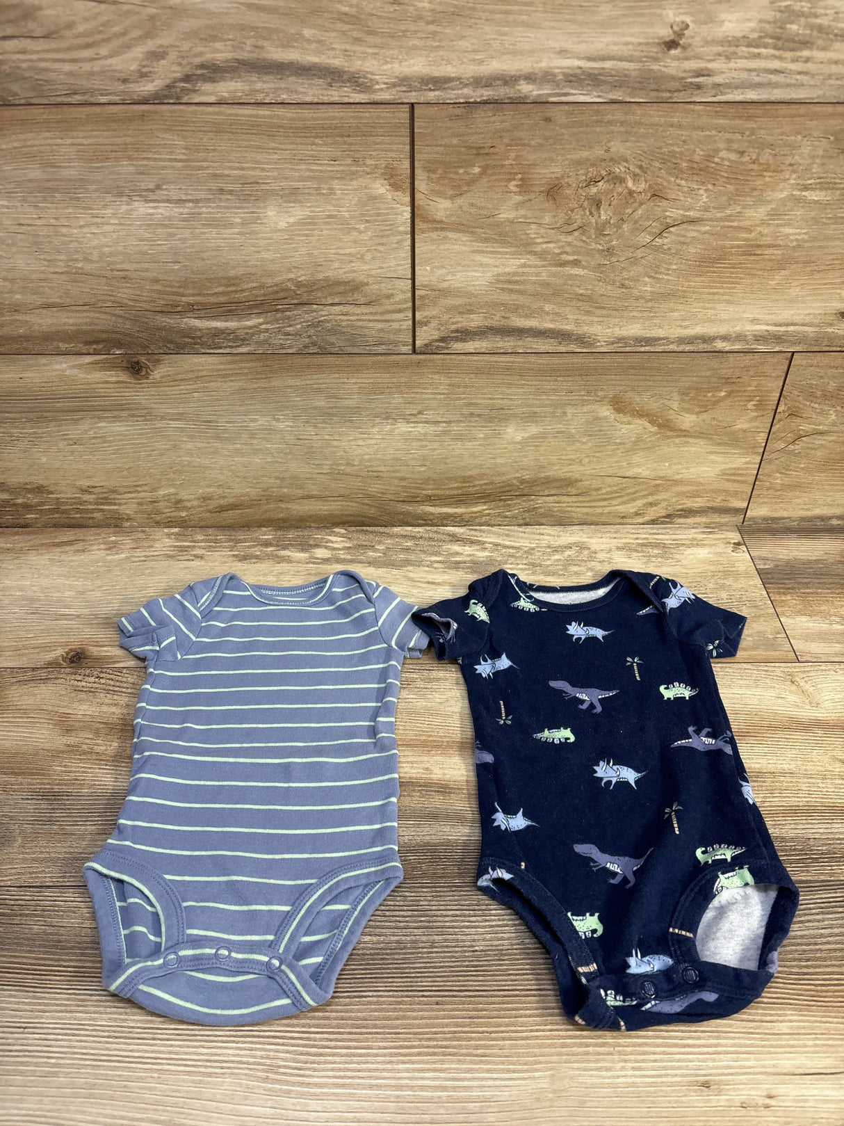 Top-down view of two Carter’s baby bodysuits: light blue with lime stripes and navy dinosaur print on wood background