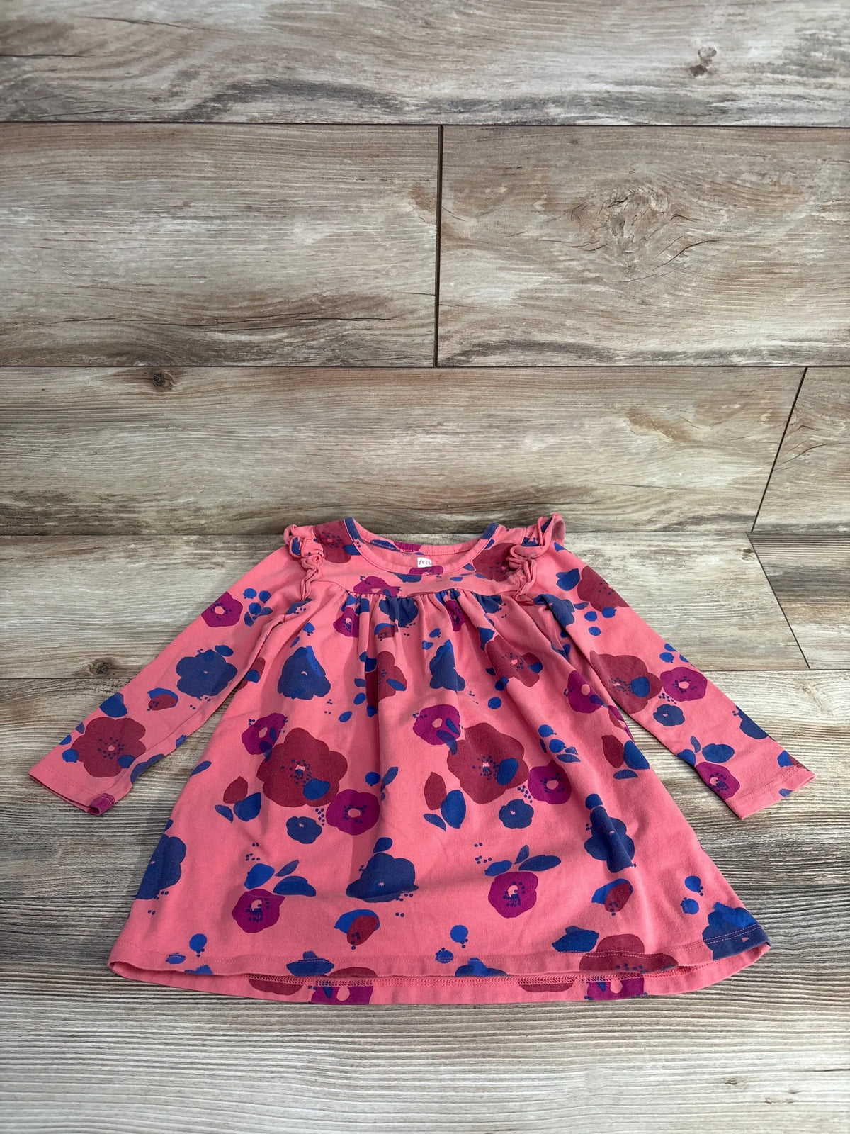 Flat lay of Tea Collection floral ruffled long-sleeve toddler dress size 18-24 months, pink with blue and purple flowers on wood floor