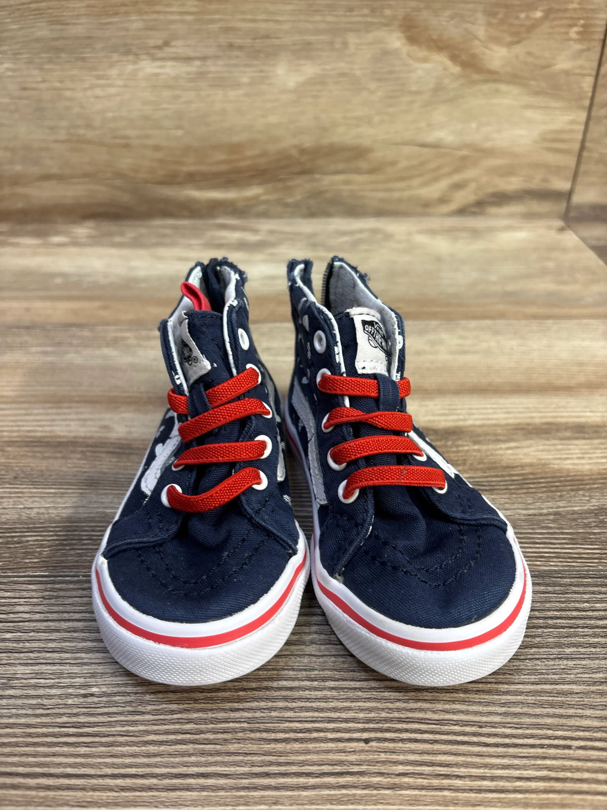 Front view of navy Vans Sk8-Hi zip toddler sneakers with red laces, white heart pattern and white rubber soles on wood background