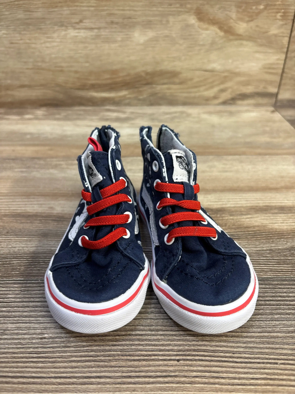 Front view of navy Vans Sk8-Hi zip toddler sneakers with red laces, white heart pattern and white rubber soles on wood background