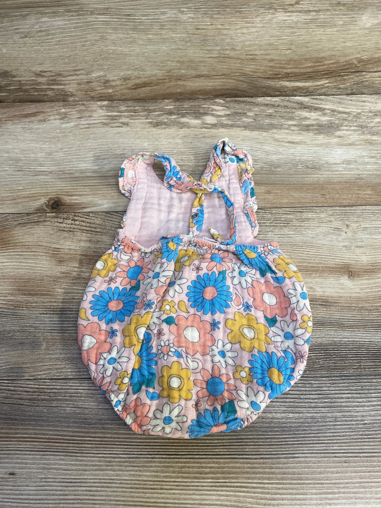 Flat lay of Angel Dear Groovy Daisy baby romper in pink with blue and yellow flowers, ruffled shoulder straps on wood background