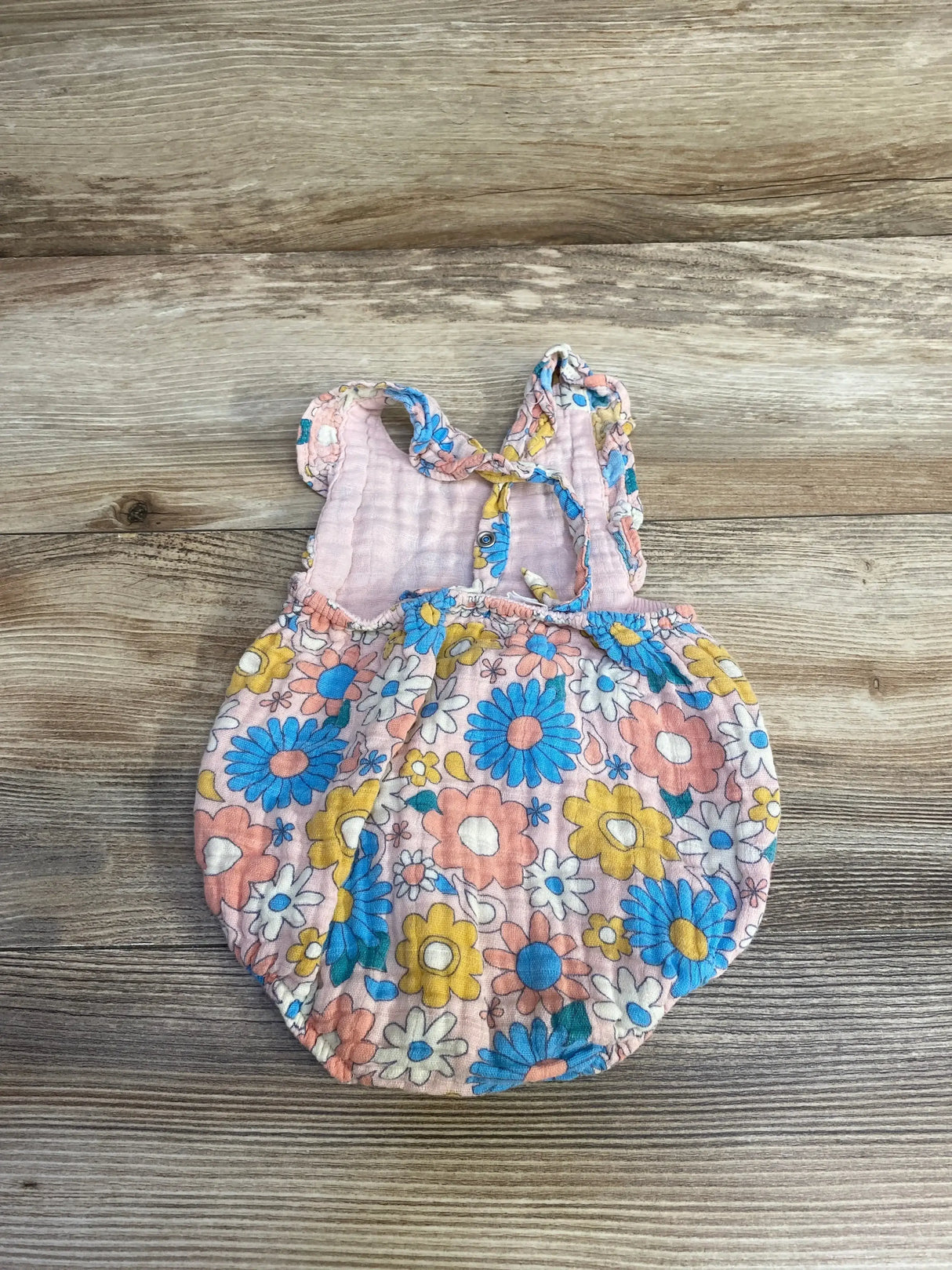 Flat lay of Angel Dear Groovy Daisy baby romper in pink with blue and yellow flowers, ruffled shoulder straps on wood background