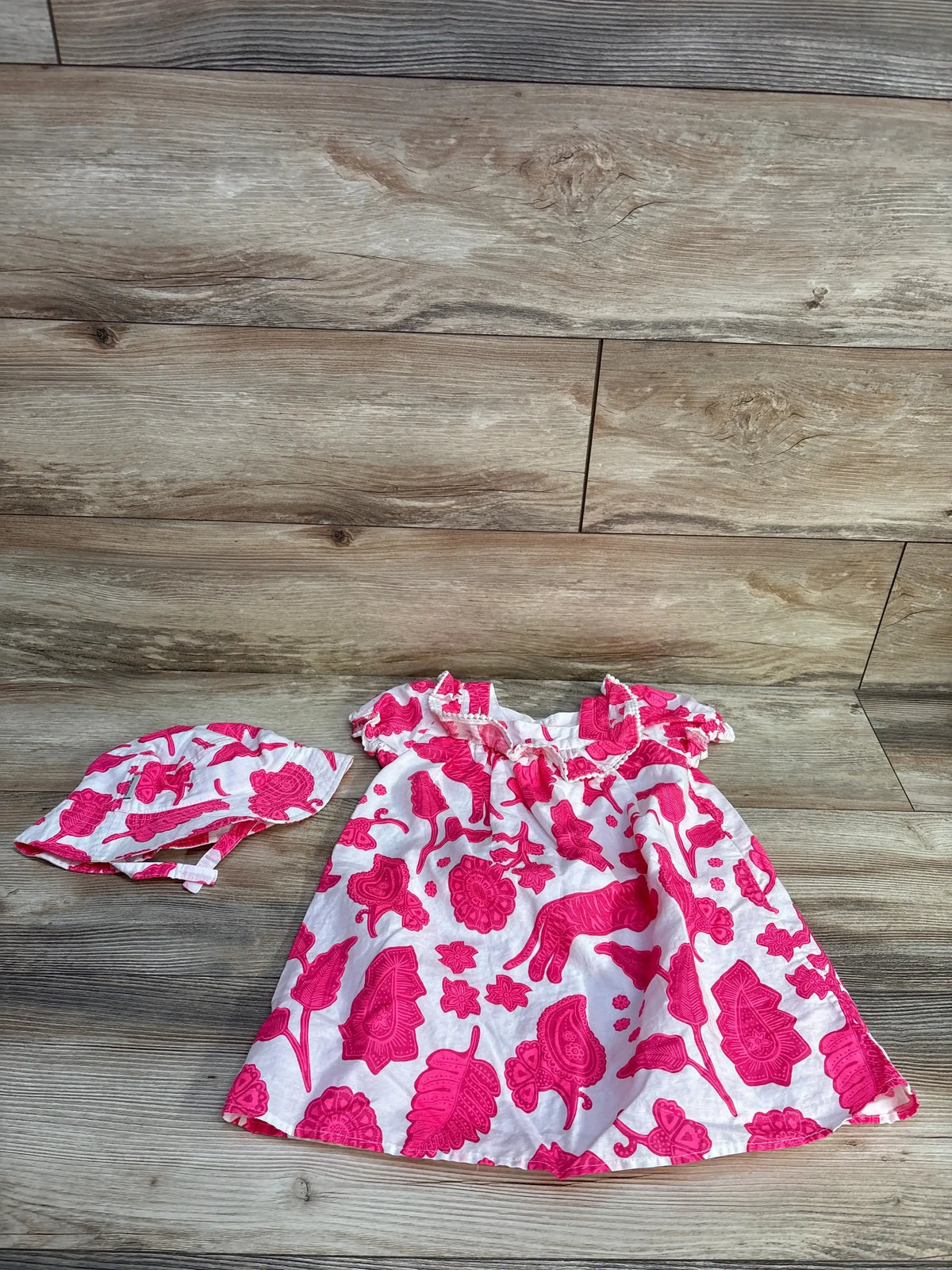 Flat-lay of white toddler linen-blend dress and matching hat showcasing bright pink floral print and ruffled neckline on wood background