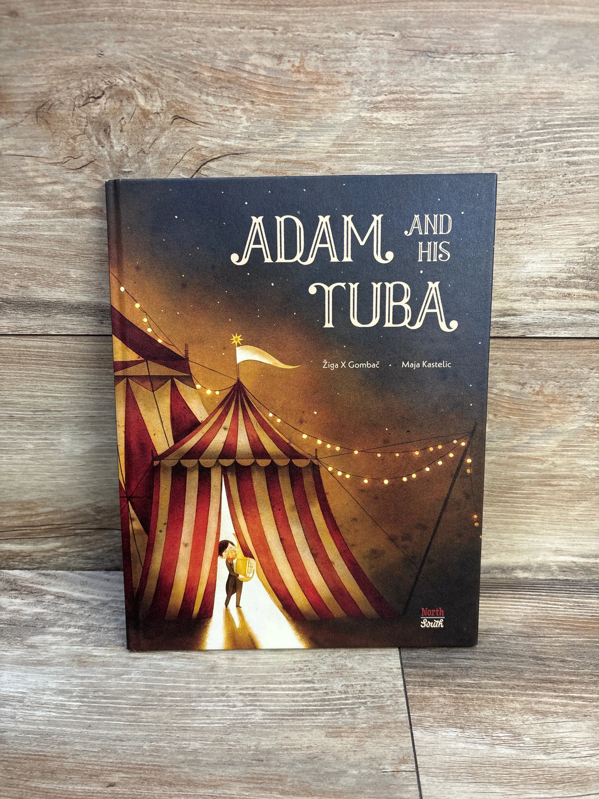 Front cover of Adam and His Tuba hardcover book showcasing a circus tent and child with a tuba on a wood-grain background