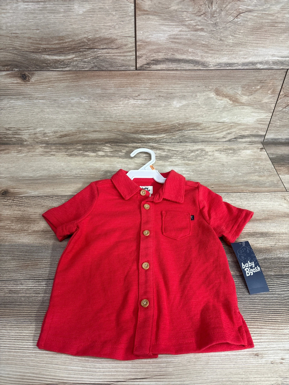Front view of red OshKosh baby button-up shirt (12 months) showcasing wooden-look buttons and chest pocket on light wood background