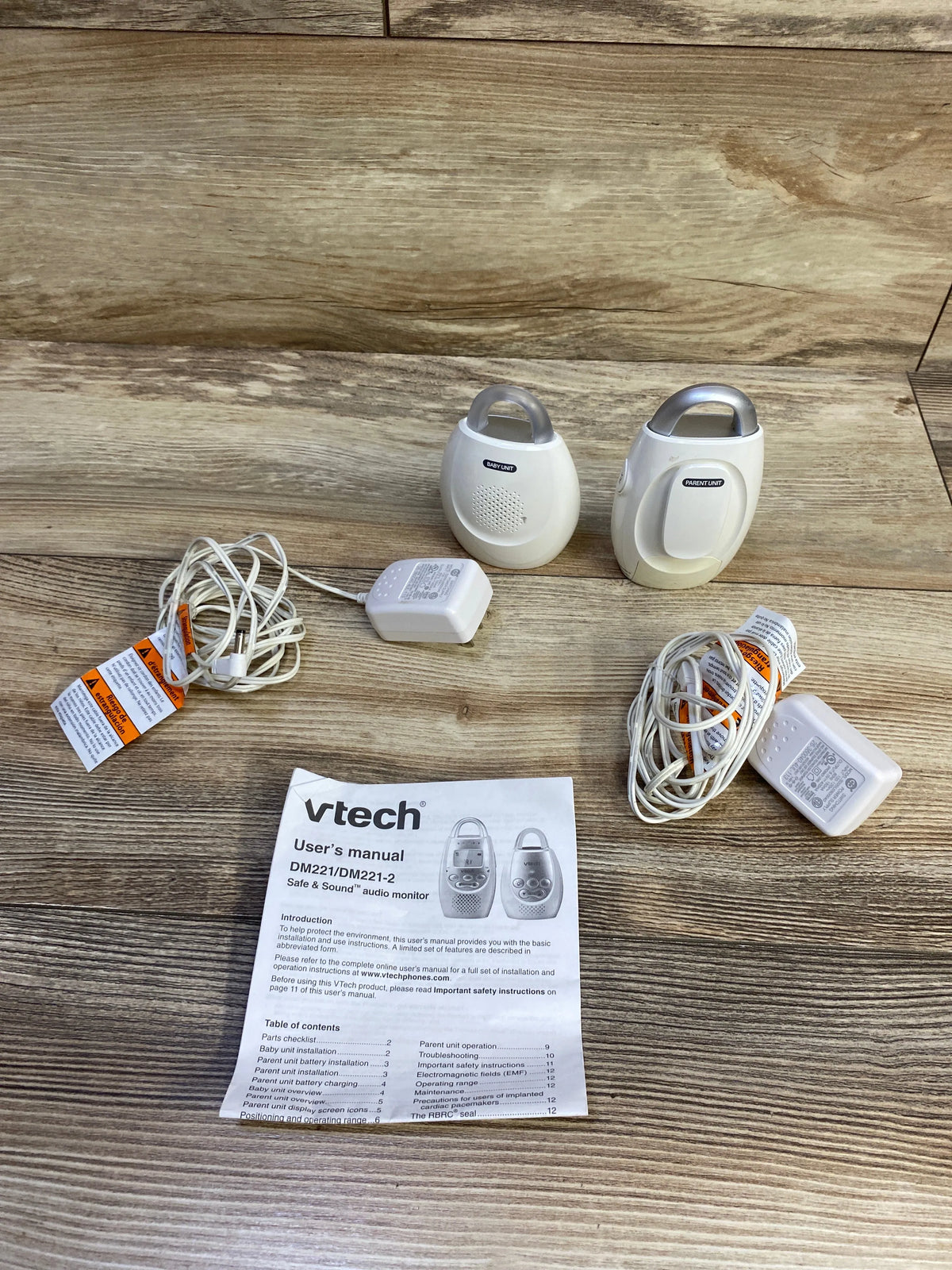 Angled top view of VTech DM221 audio baby monitor showing parent and baby units, two power adapters, and user manual on a wood floor