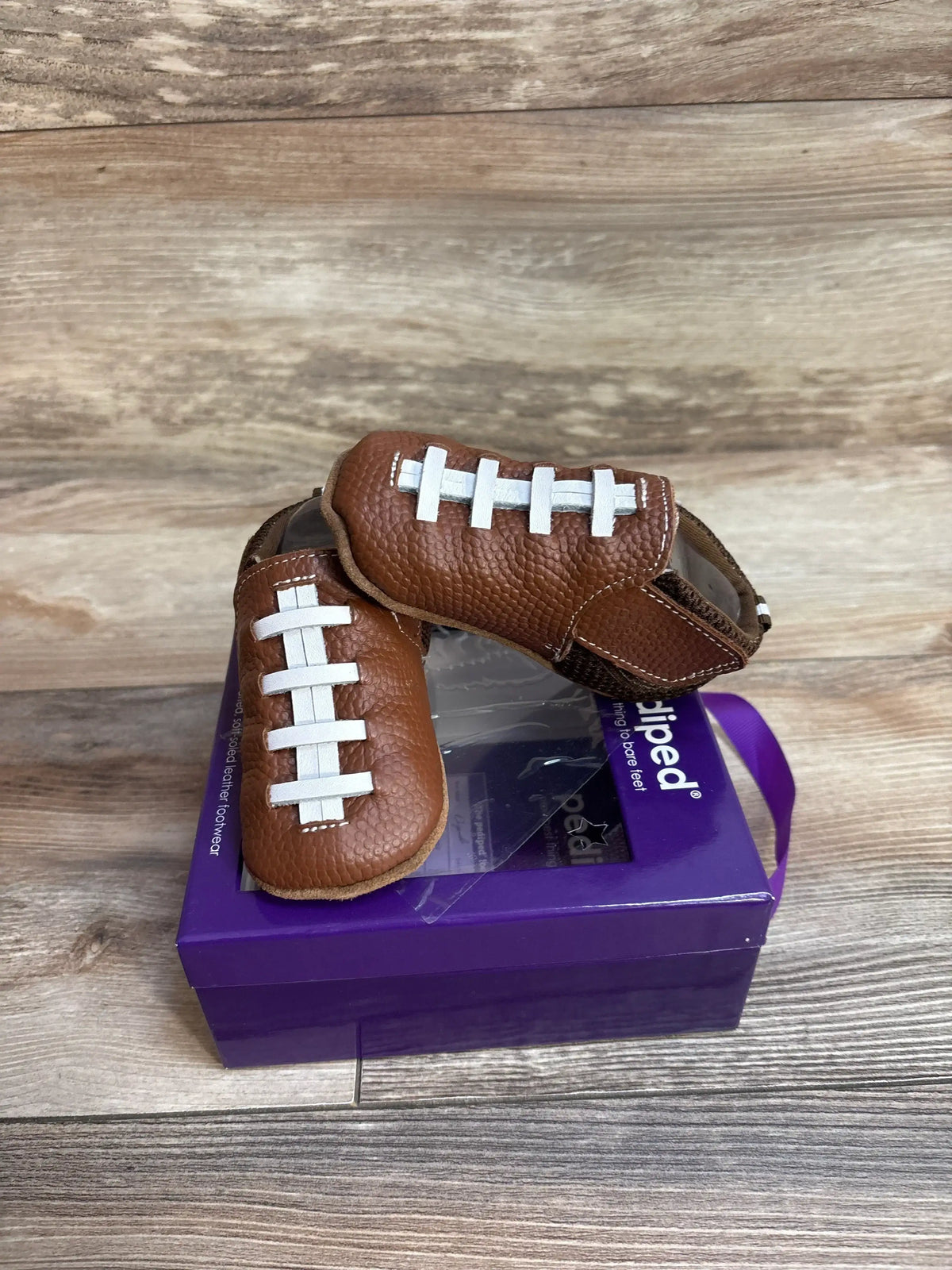Brown leather baby shoes designed like footballs on a purple pediped box on wooden floor