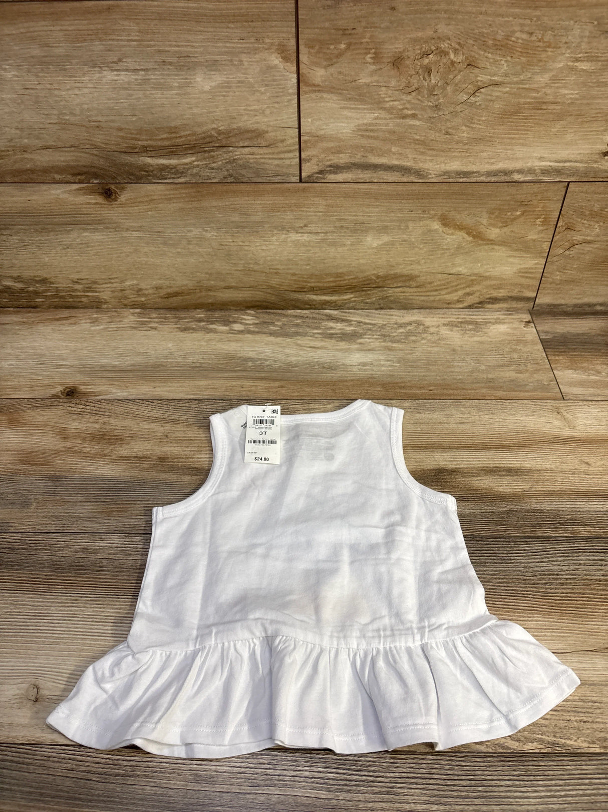 Flat lay of white Epic Threads peplum tank top showing ruffled hem and visible size 3T tag on wood floor background