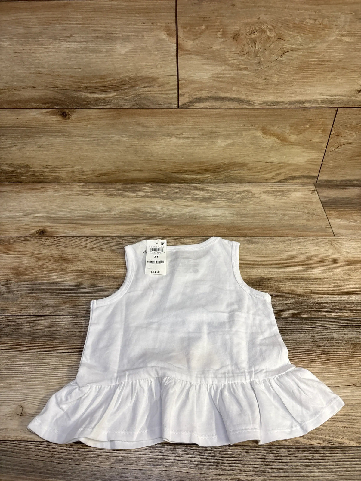 Flat lay of white Epic Threads peplum tank top showing ruffled hem and visible size 3T tag on wood floor background