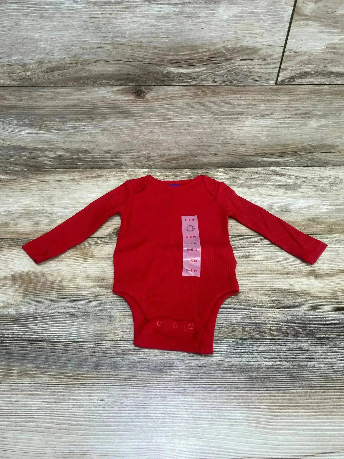 Flat lay of red rib-knit long-sleeve baby bodysuit with 3 snap crotch and pink size tag 3-6M on wood background