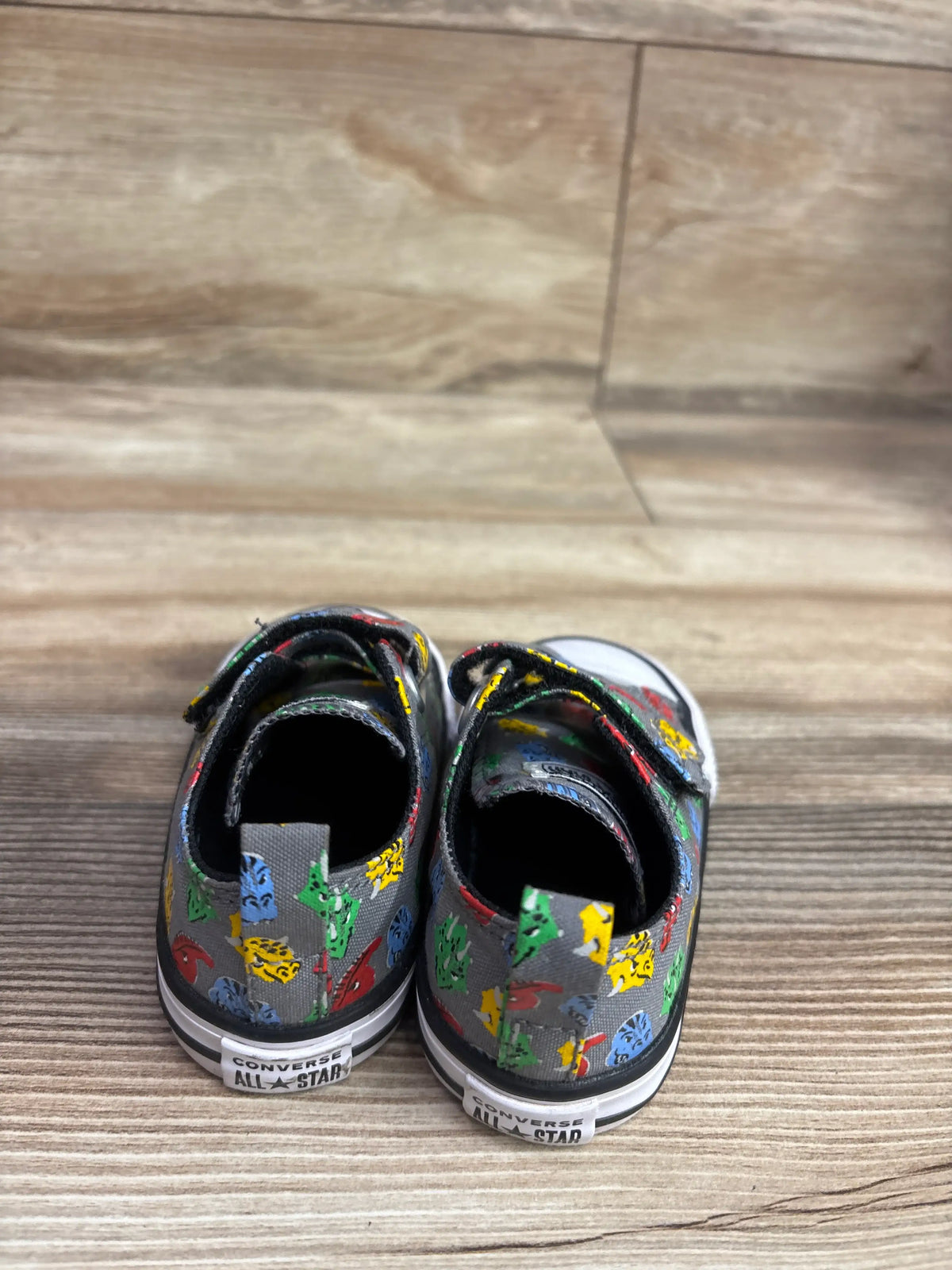 Rear view of Converse Chuck Taylor All Star 2V Dino sneakers showcasing colorful dinosaur prints and All Star heel branding on a wood floor