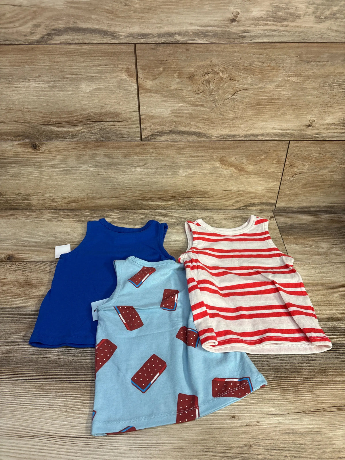 Top view of Cat & Jack 3pk ice cream tank tops showcasing blue, ice cream print, and red striped designs on a wood floor