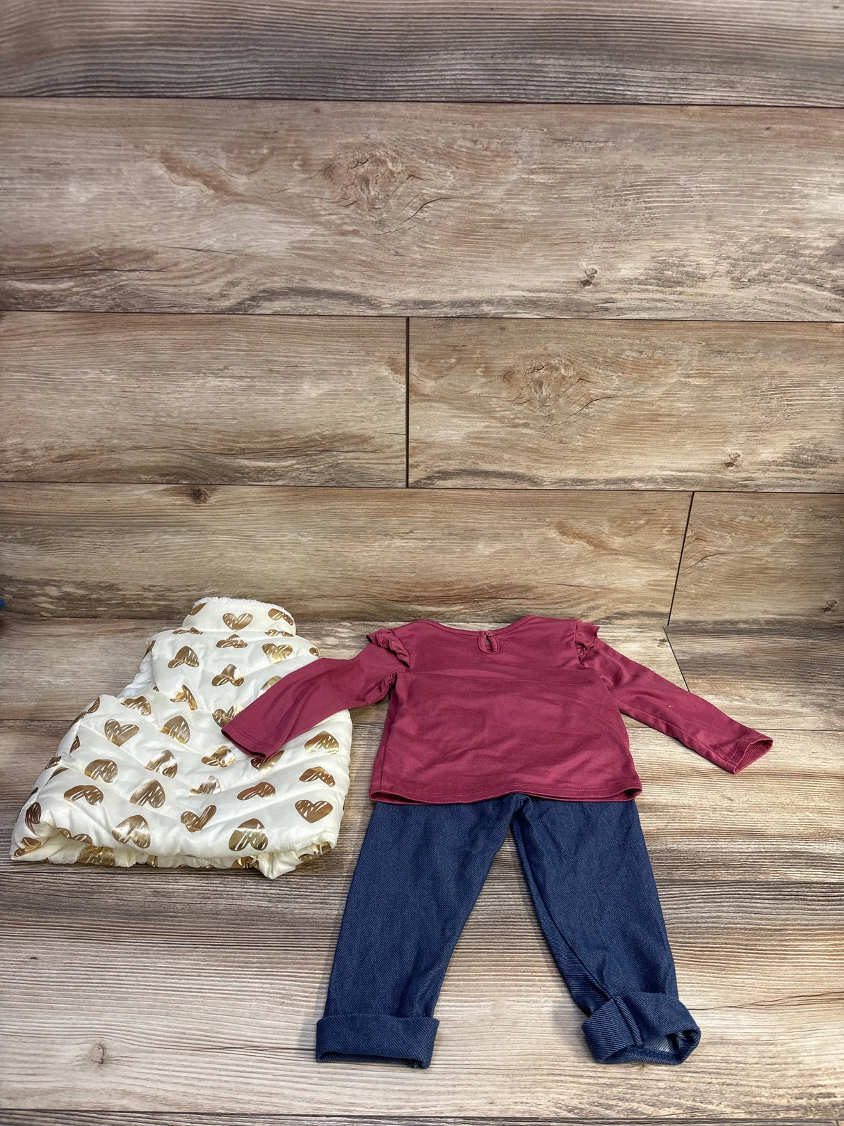 Flat lay of toddler 3-piece set: white heart-print puffer vest, pink long-sleeve ruffle top, cuffed navy jeggings on wood