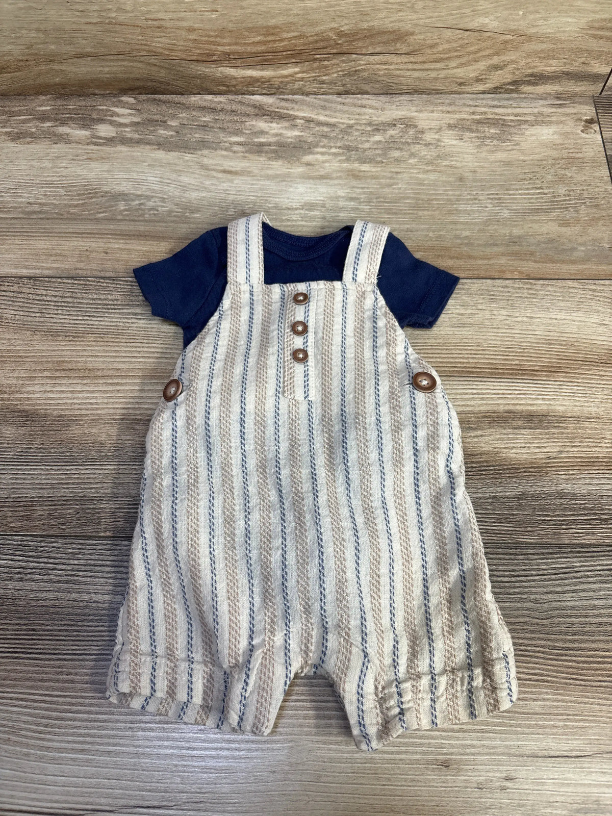 Front view of Just One You 2pc bodysuit and shortall set showing beige and blue stripes, navy bodysuit, and buttons on wood