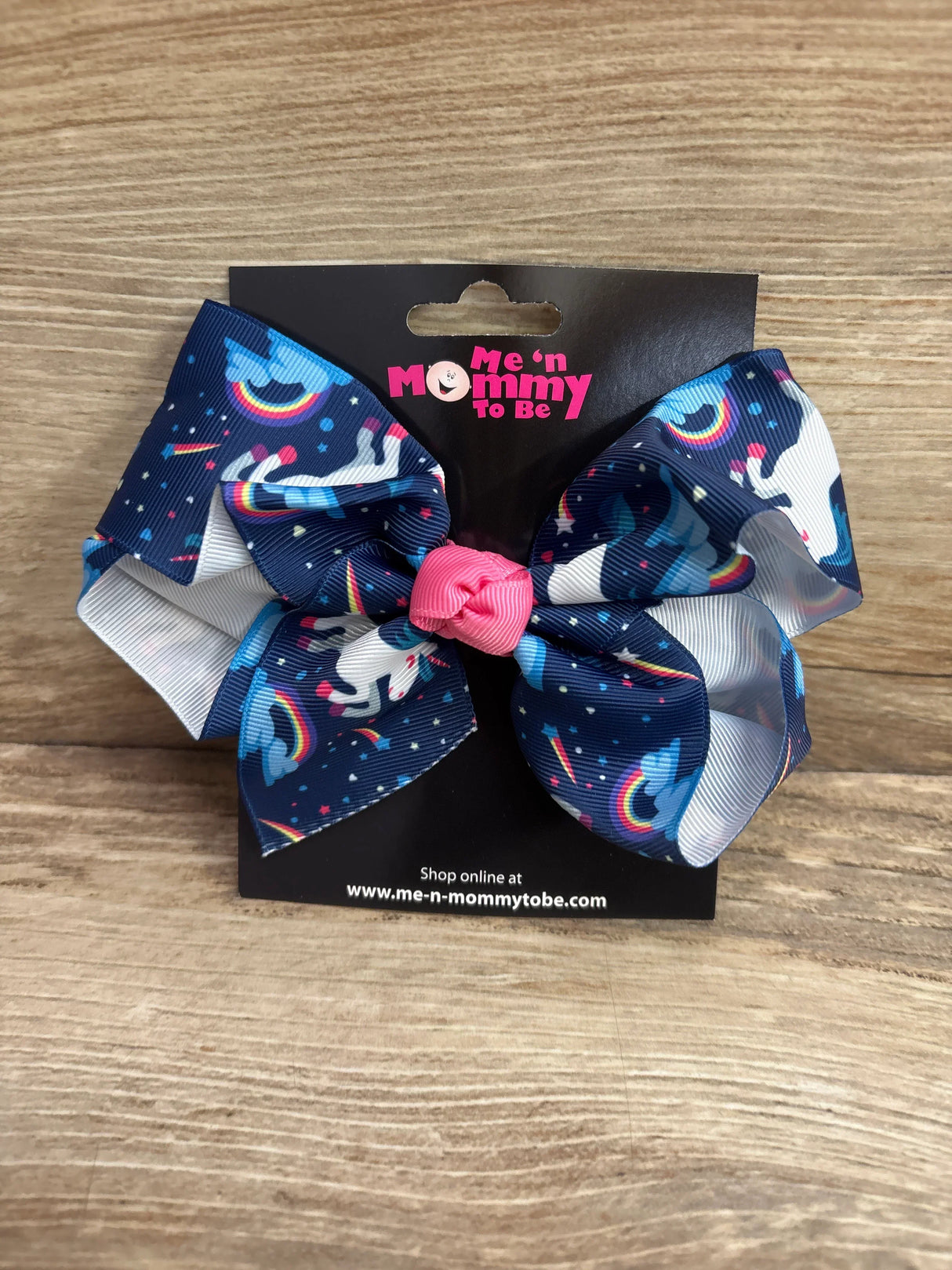 Front view of large navy unicorn hair bow with rainbow print and pink center knot on black branded card on wood background