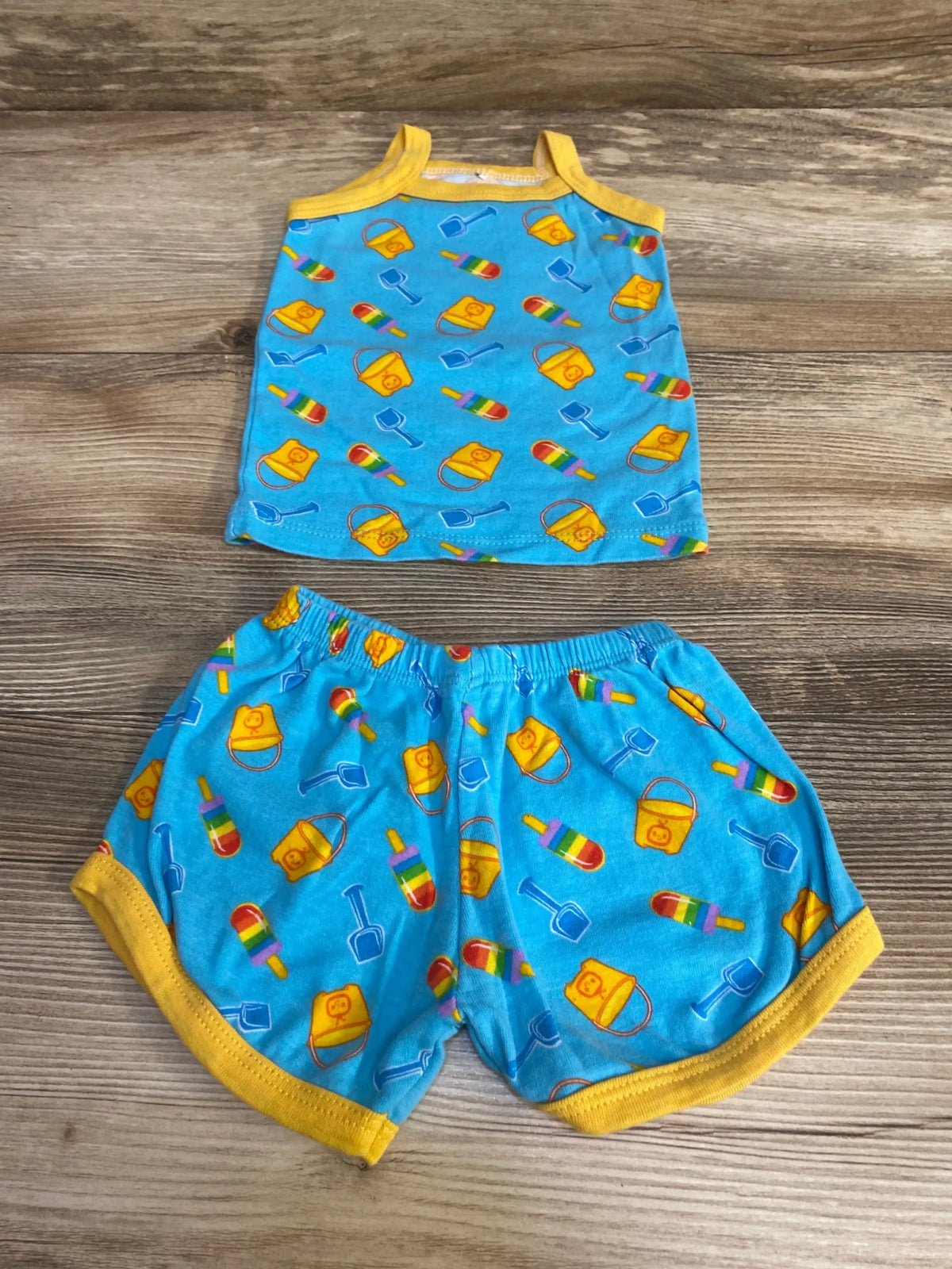 Flat lay of newborn 2-piece Cocomelon tank top and shorts showcasing yellow trim and bucket, shovel and popsicle print on wood background