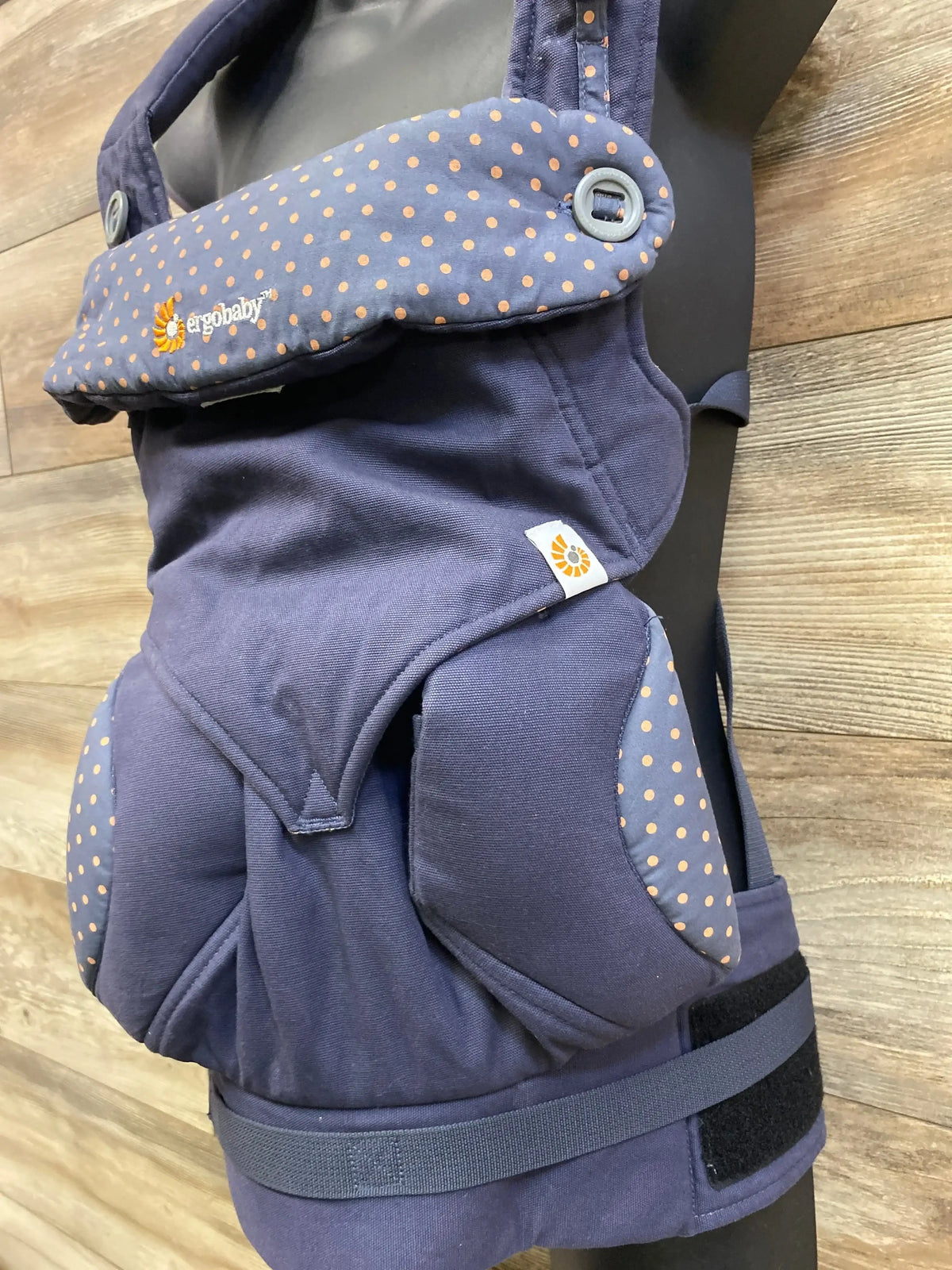 Three-quarter view of Ergobaby 360 baby carrier in dusty blue with orange polka dot trim and 12 to 33lbs capacity on wood plank background