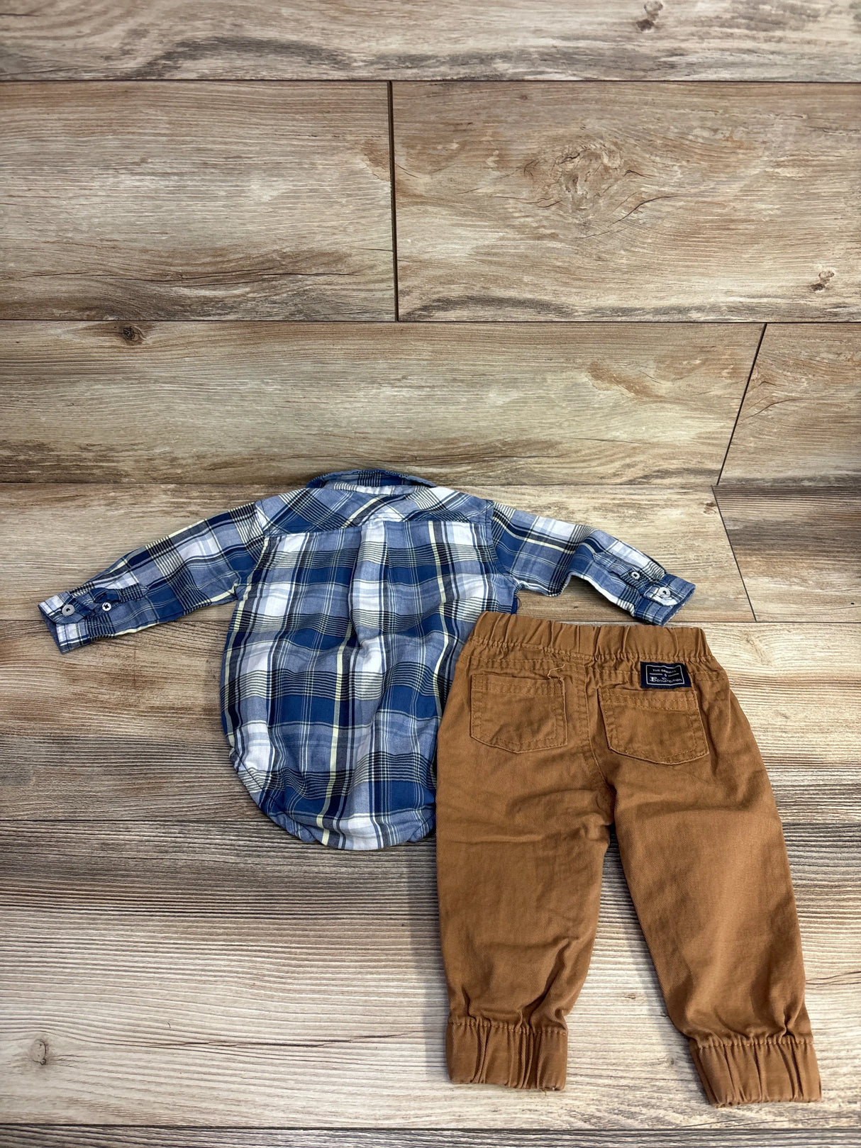 Children's blue plaid button-up shirt and brown elastic cuff pants laid flat on wooden floor