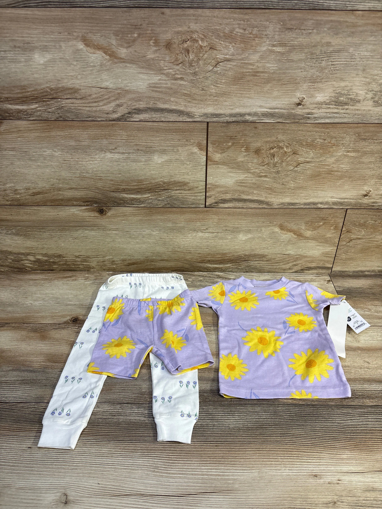 Flat lay of Just One You 3-piece floral pajama set showcasing lavender top and shorts with large yellow sunflowers and white floral leggings on wood floor