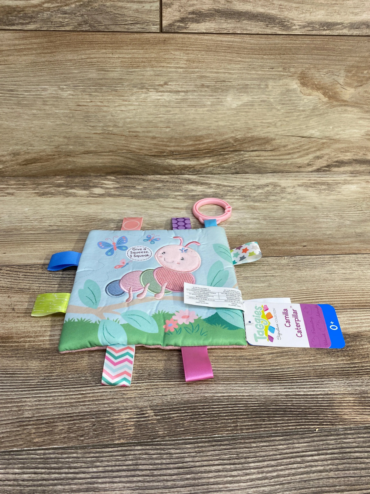 Taggies Camilla Caterpillar soft baby sensory toy with tags, flowers, and ring clip on wood background