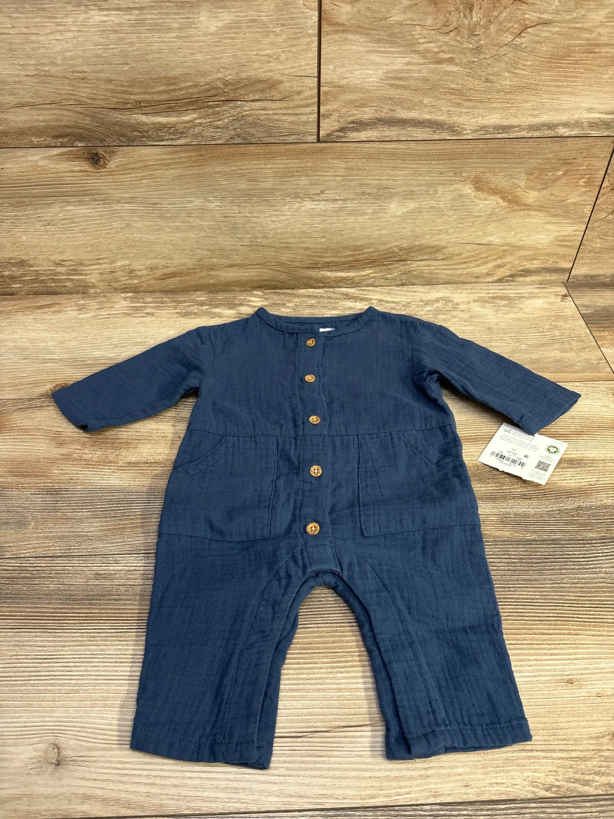 Front flat-lay of blue Little Planet muslin baby jumpsuit with wooden buttons, long sleeves and pockets on wood surface