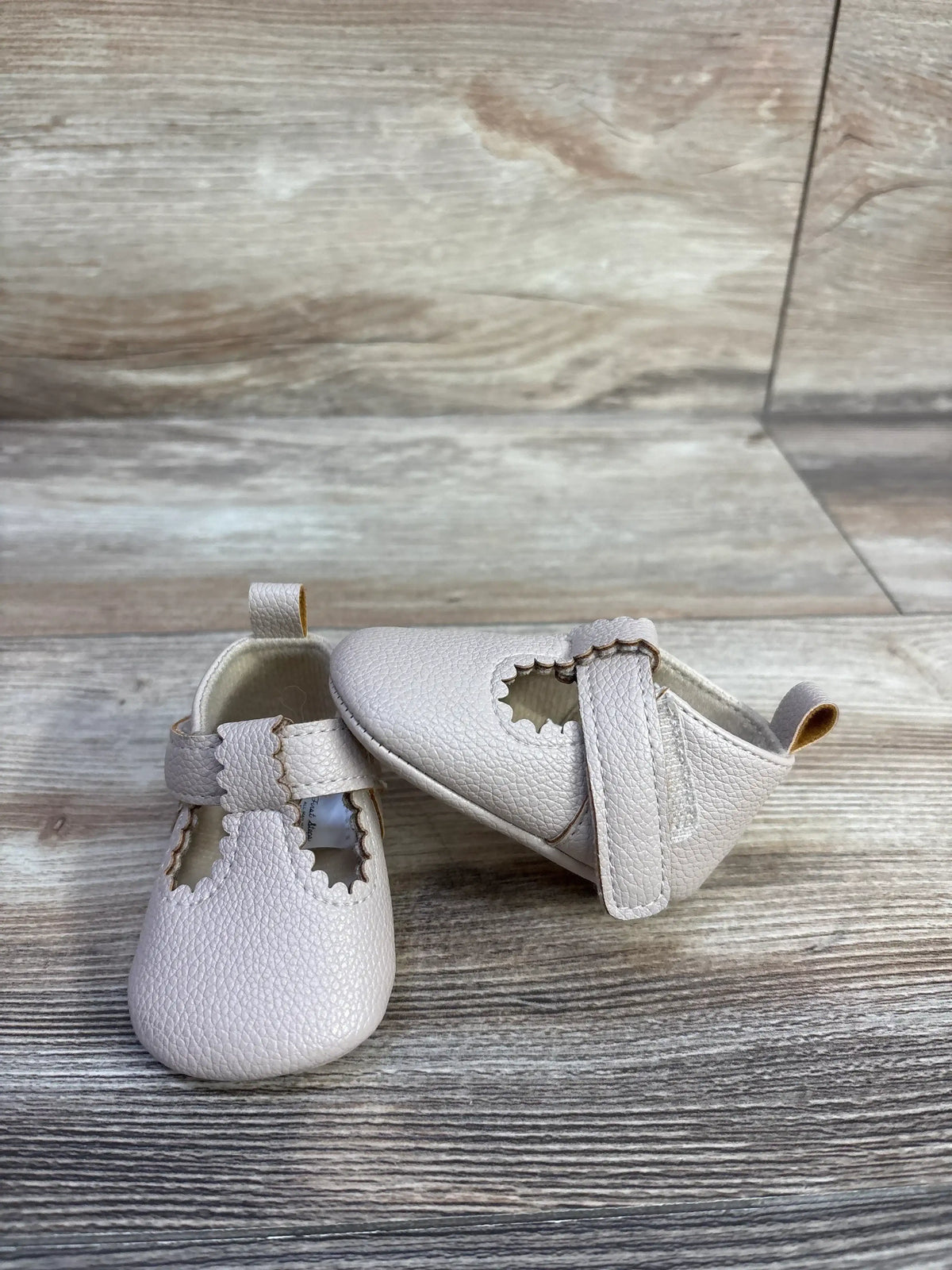 Front and side view of First Steps soft sole baby shoes showcasing scalloped T-strap and pebbled beige leather on light wood background