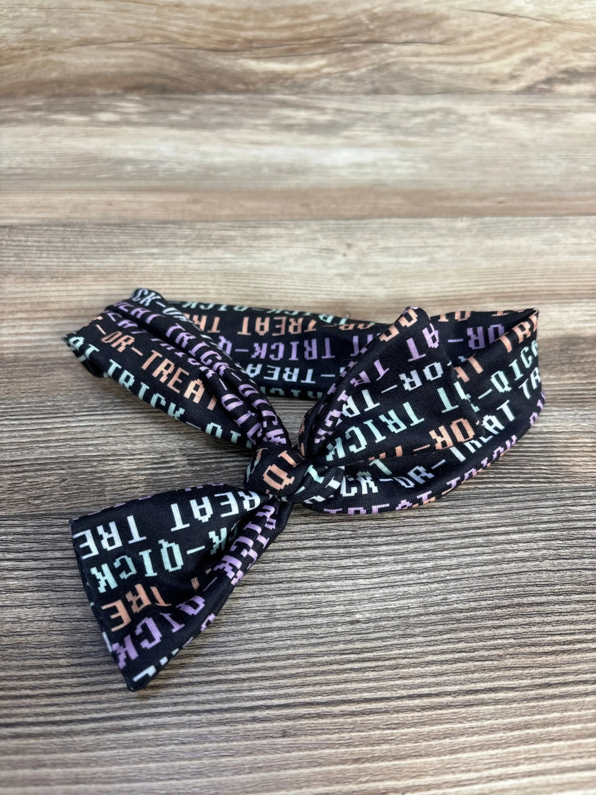Angled view of one-size Halloween headwrap showcasing a knotted bow and multicolor Trick-or-Treat print on black fabric