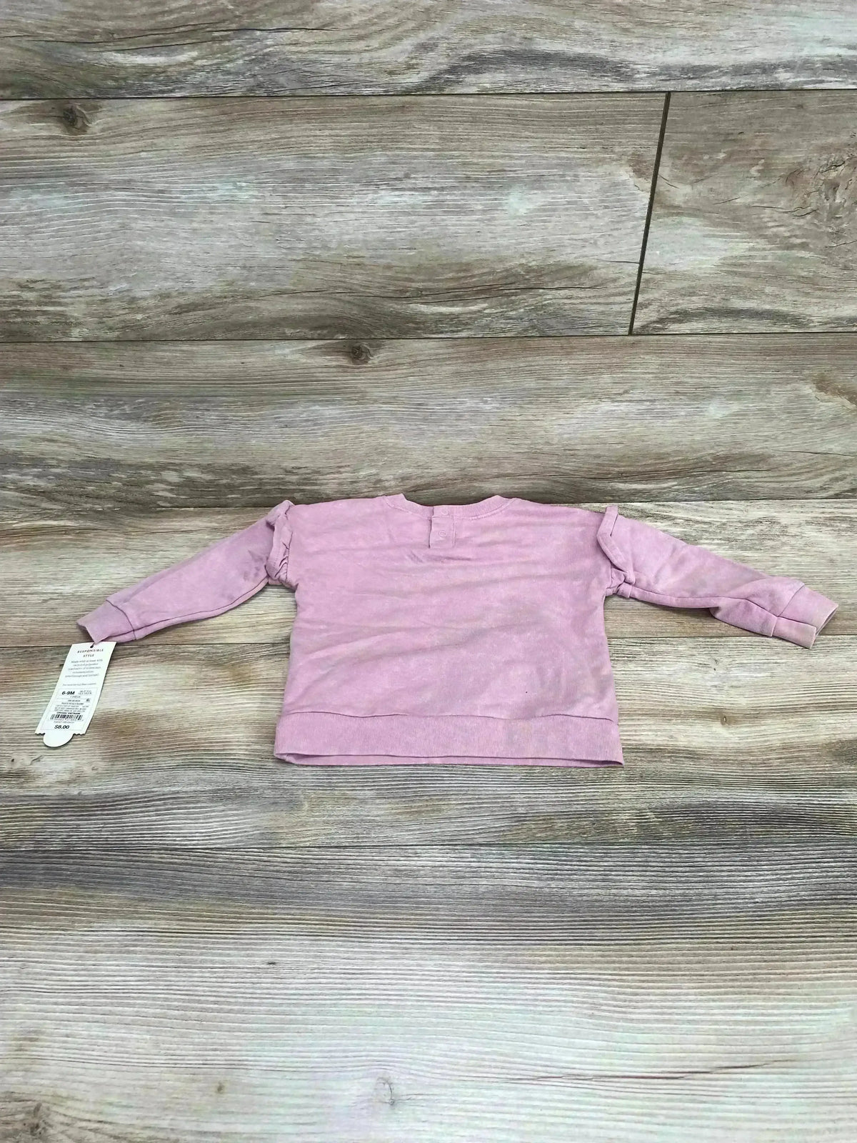 Flat lay of pink Cat & Jack baby sweatshirt, size 6–9 months, showing long sleeves, ruffle shoulder detail and back snap on wood background
