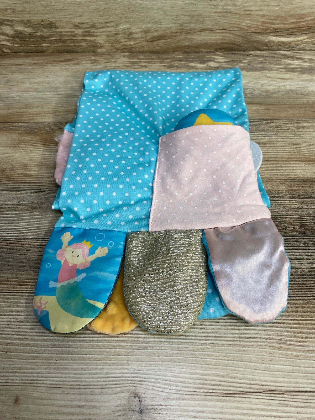 Top-down view of Demdaco mermaid activity scarf with blue polka-dot fabric, pink pocket, and shimmery tail flaps on wood