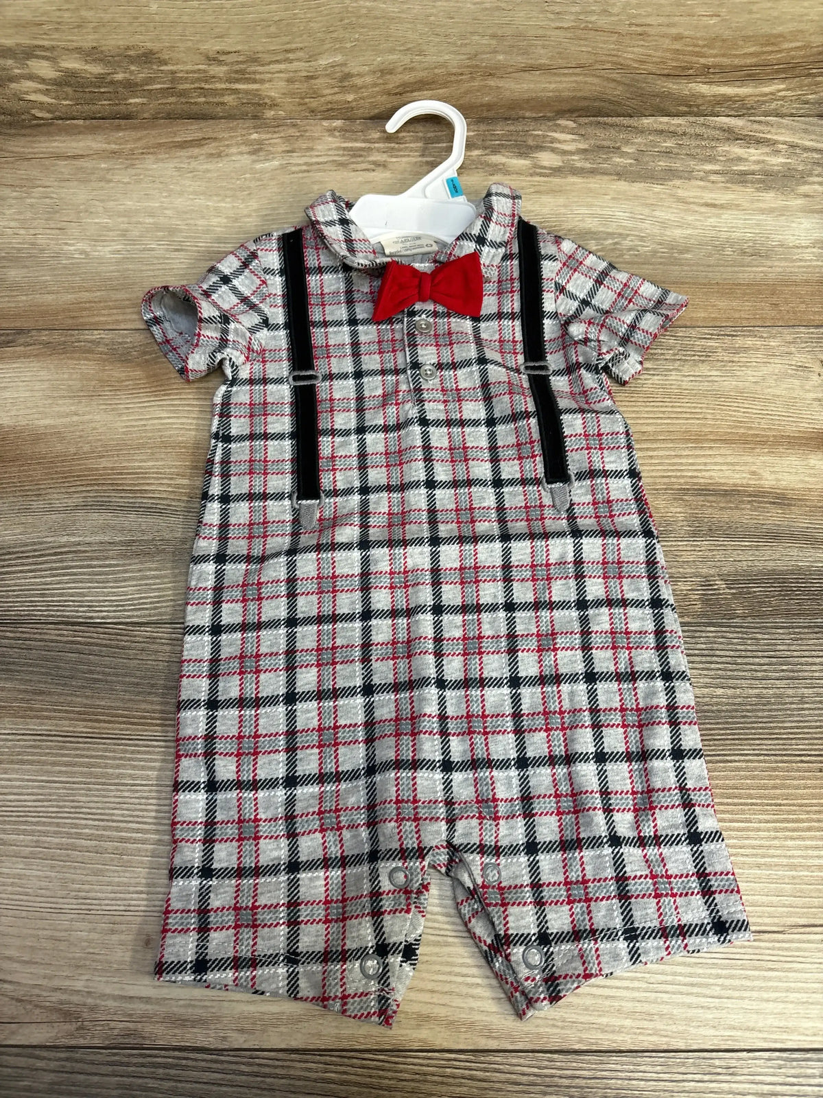 Front view of grey plaid baby romper 3–6m with red bow tie, black faux suspenders, short sleeves, snaps at legs on wood
