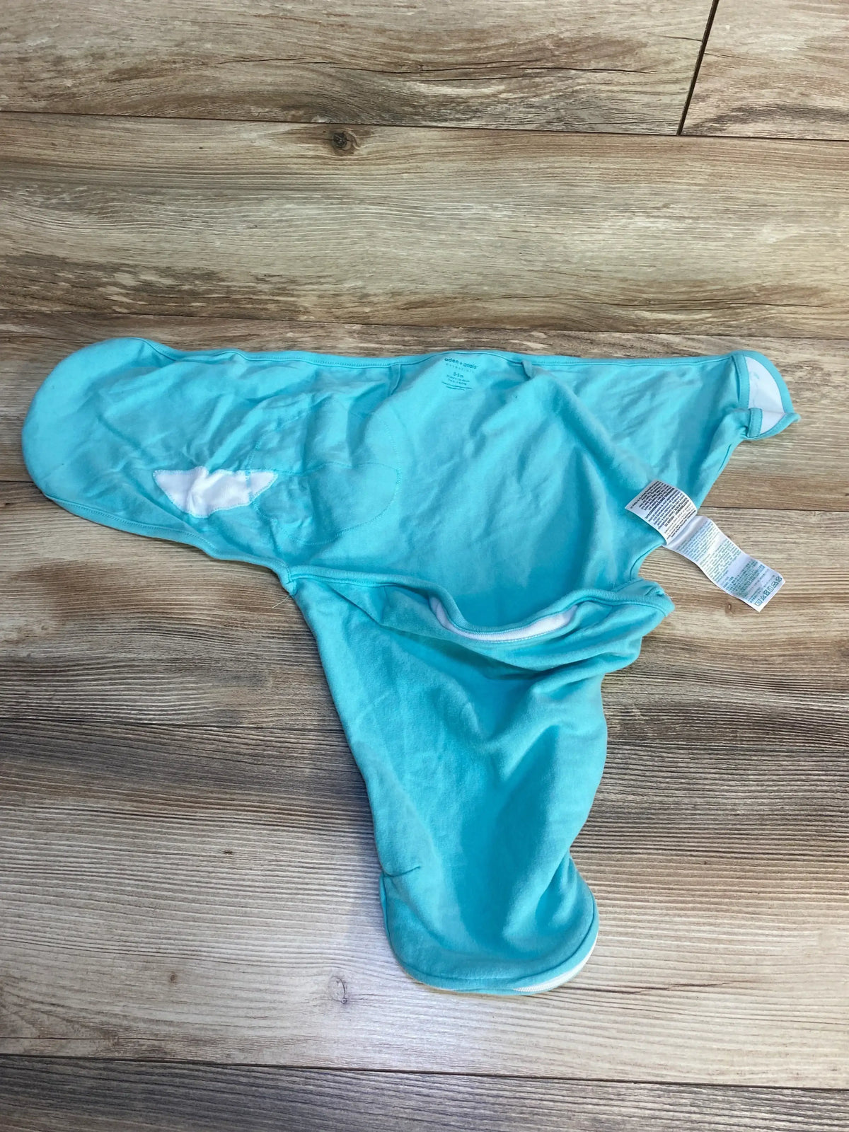 Top-down view of Aden + Anais newborn swaddle wrap showcasing zip closure, winged ergonomic shape, and white interior lining with care tag visible on wood floor