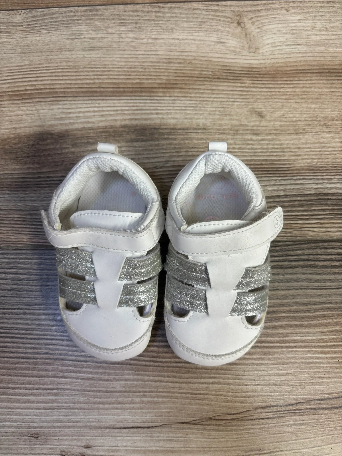 Top view of Surprize by Stride Rite white fisherman sandals with silver glitter straps on a wood-grain background