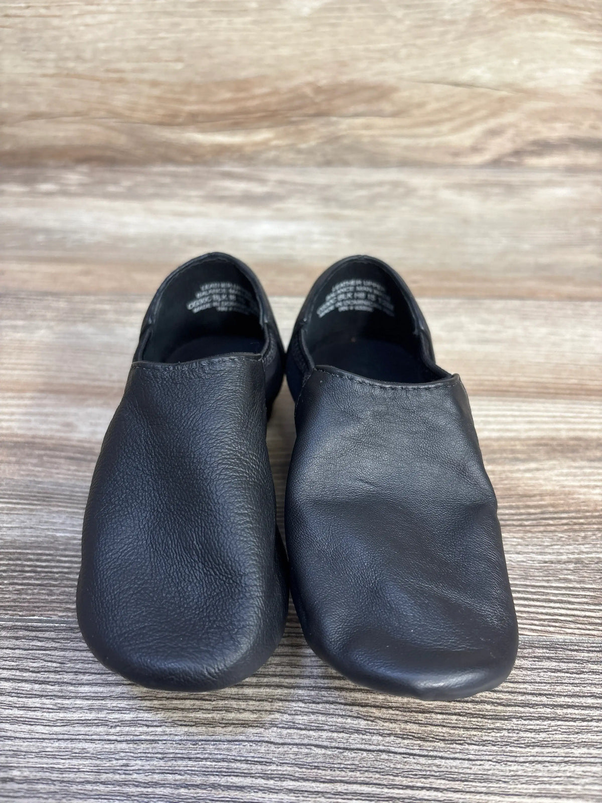Front view of black Capezio E-Series Jazz slip-on shoes showcasing soft leather uppers and low sides on a wood background