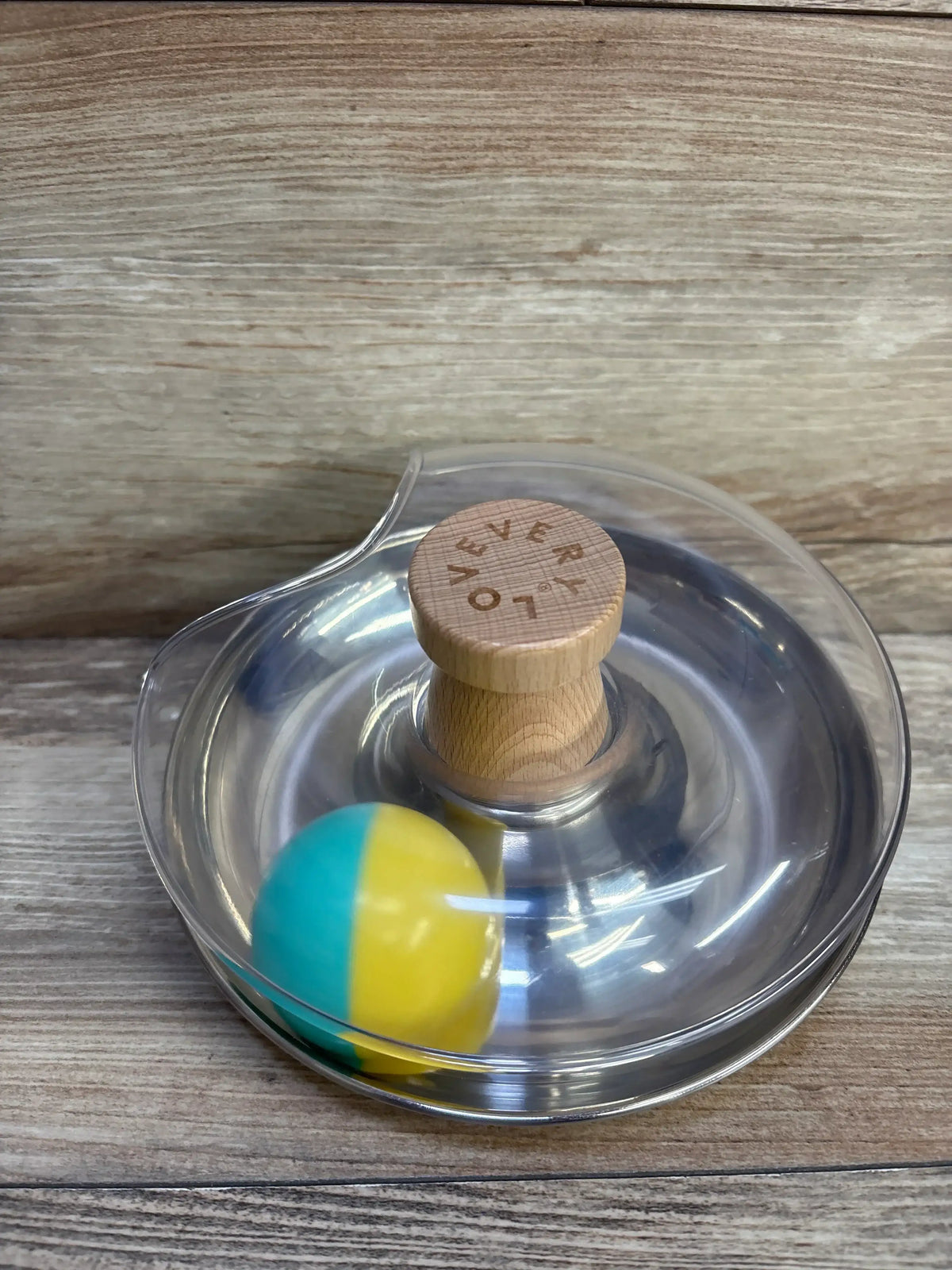Top view of Lovevery Tip & Turn toy showing stainless steel base, clear dome, wooden knob with logo and yellow-teal ball