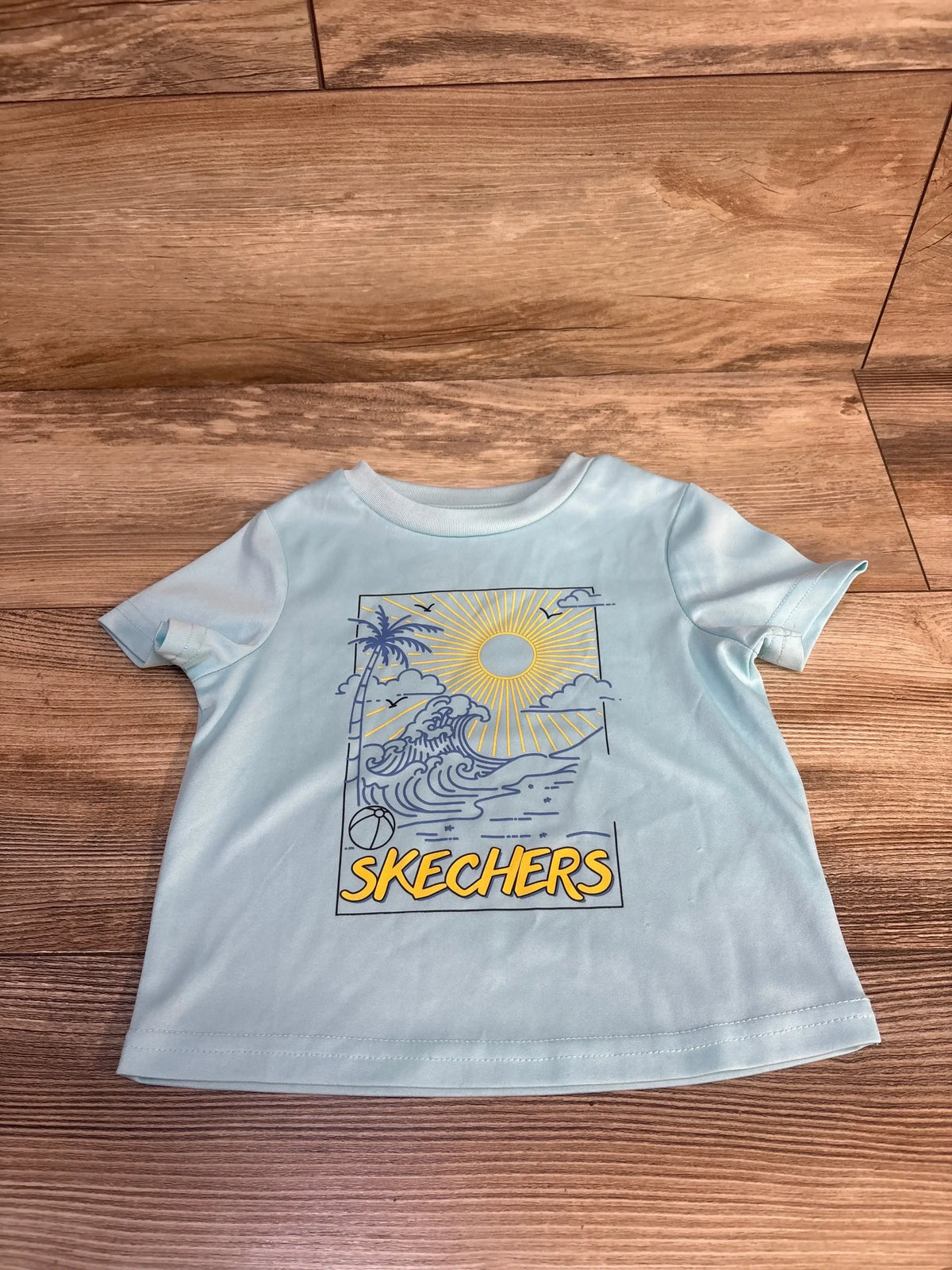 Light blue kids Skechers t-shirt with beach graphic, sun, palm tree, waves, and beach ball on wood floor