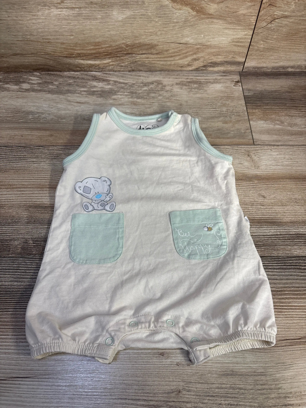 Front view of cream newborn tank shortie romper showing mint trim, two front patch pockets, bear graphic, and snap crotch on wood floor