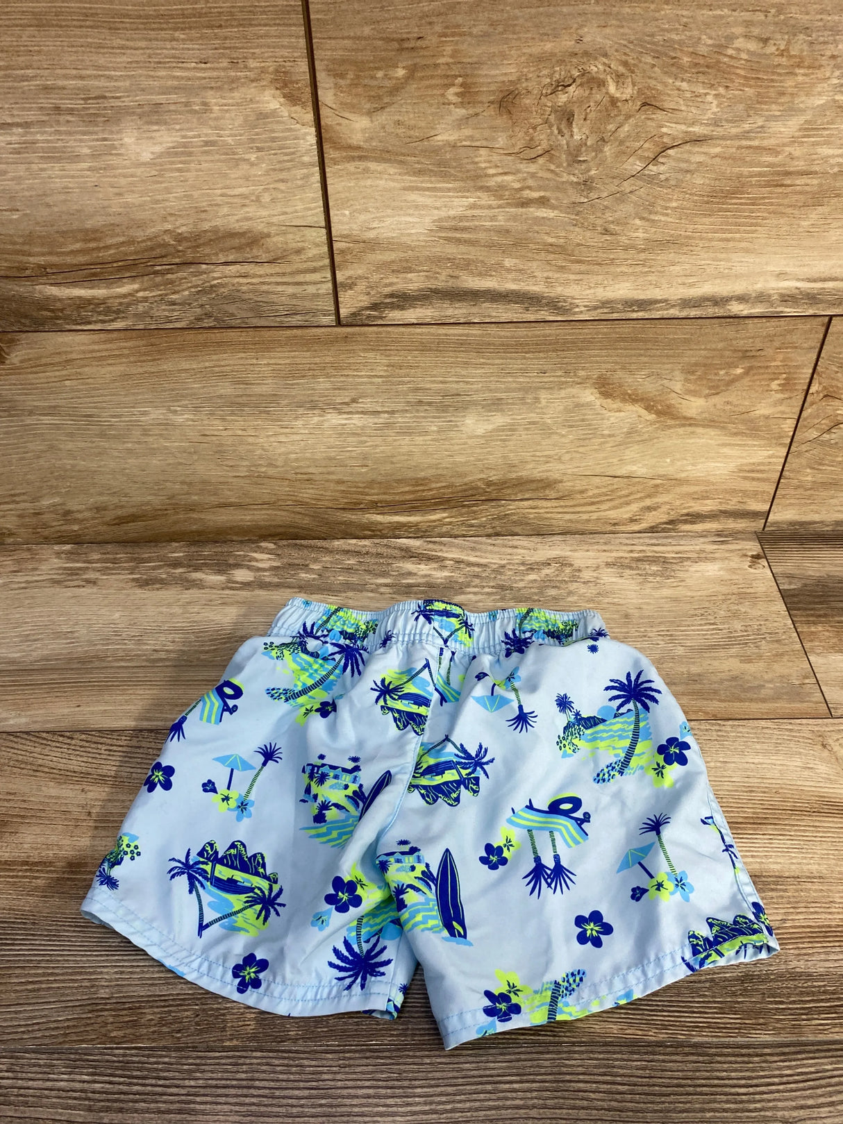 Front view of Cat & Jack swim trunks 4T, light blue with navy and lime tropical print, elastic waistband, on wood background