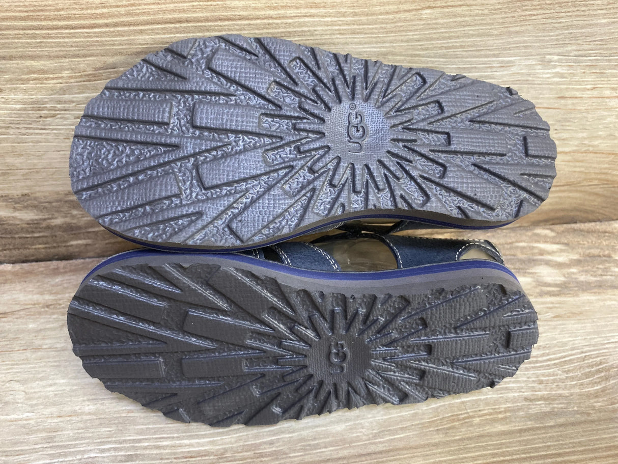 Bottom view of UGG Josiah toddler fisherman sandals showcasing navy rubber tread pattern and UGG logo on wood background