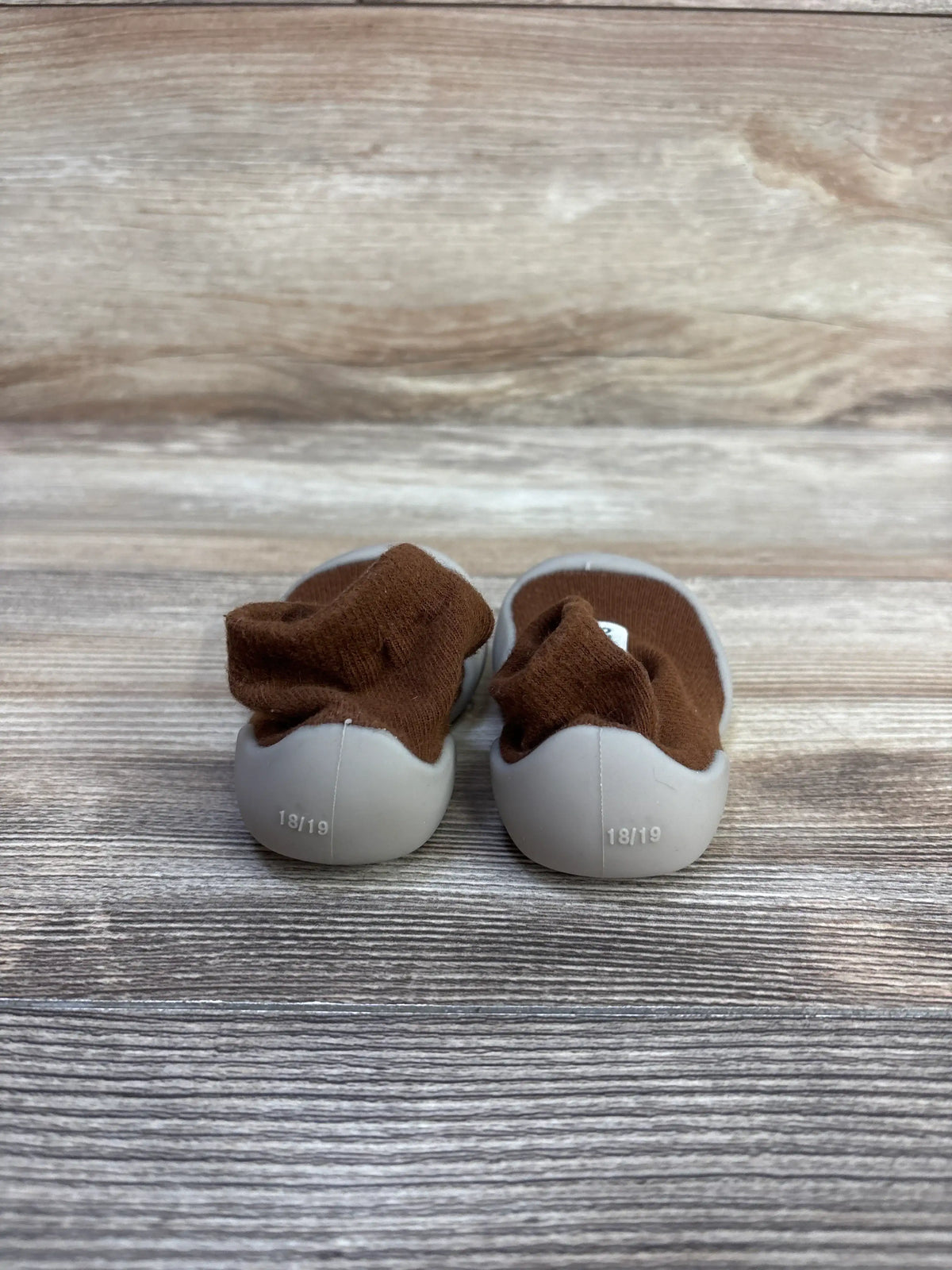 Rear view of Tiny Explorings Self-Care Sock Shoes Brown sz 2/3c showcasing light gray soles marked 18/19 on a wood-grain background