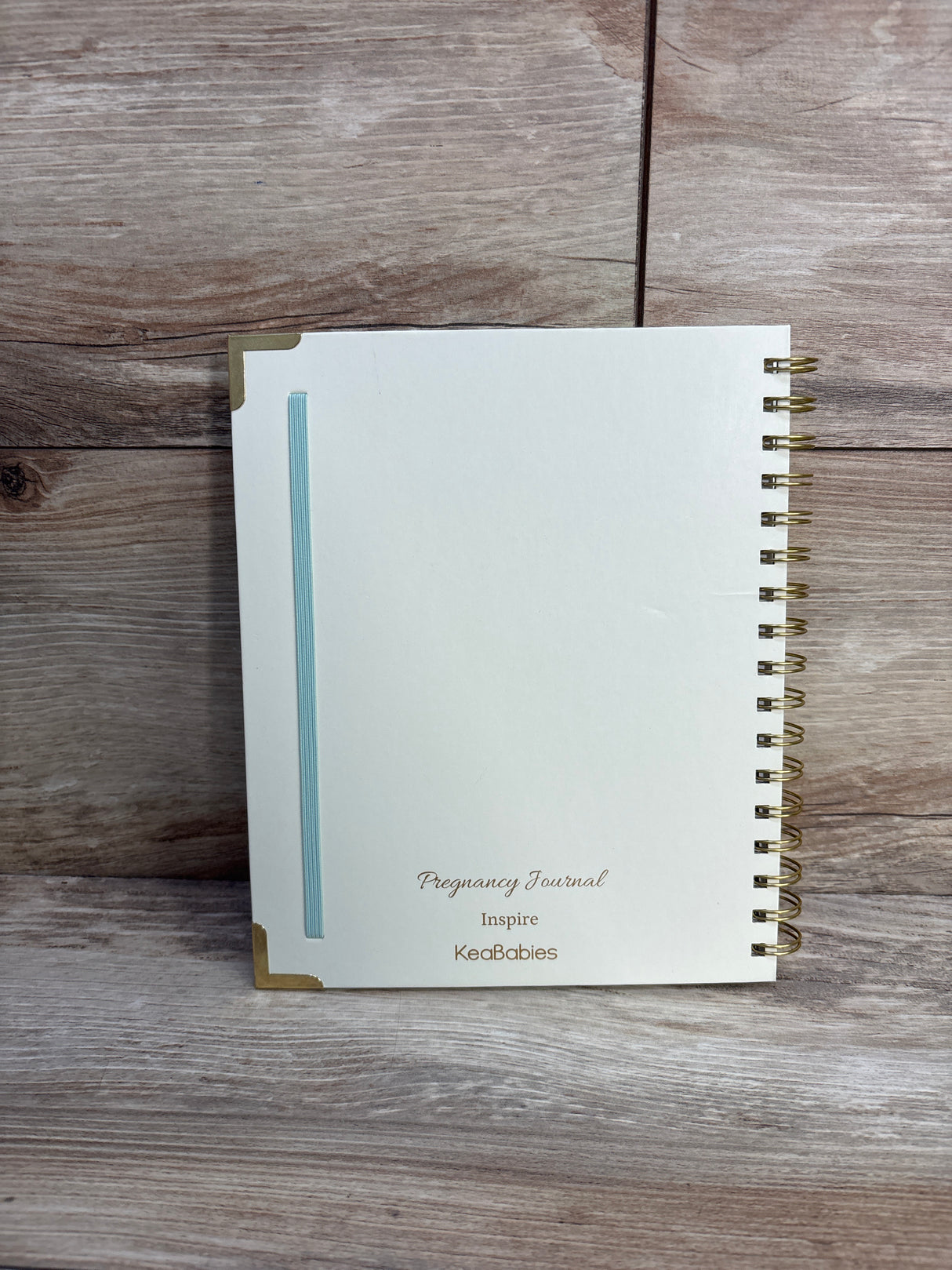 Front view of KeaBabies Inspire Pregnancy Journal showcasing a light blue hardcover, gold spiral binding, and elastic closure