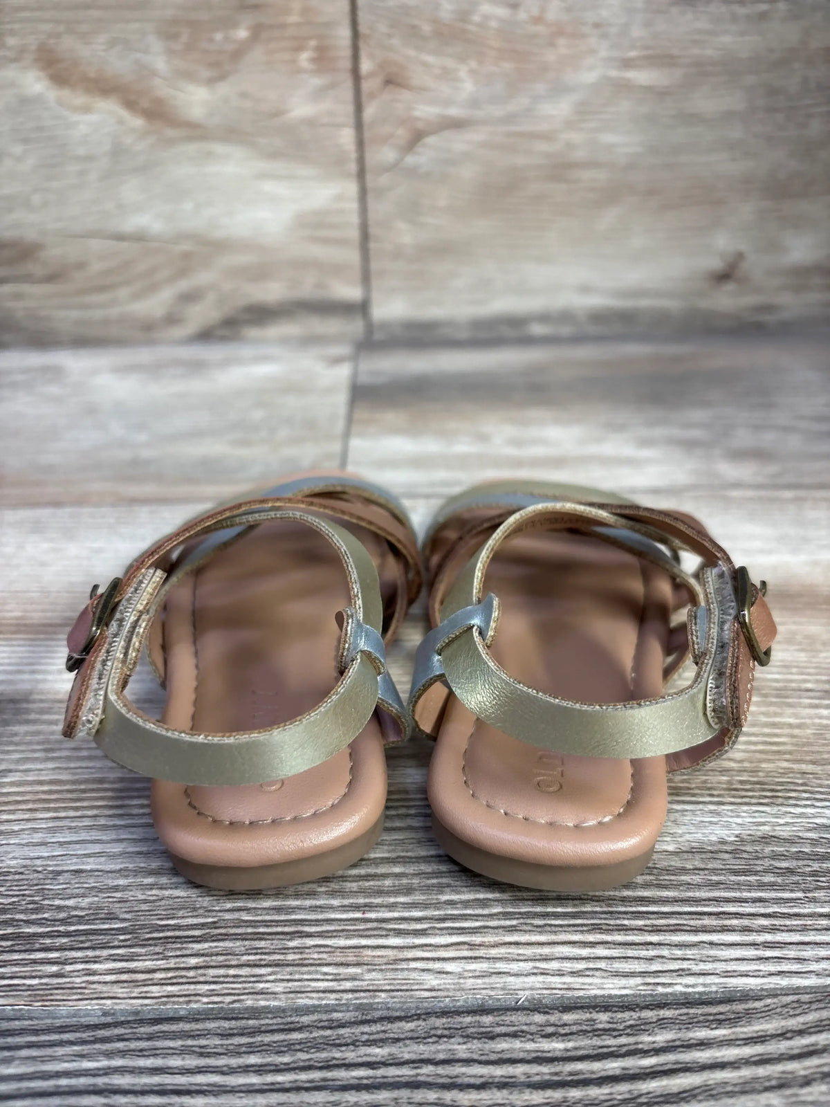 Rear view of toddler metallic gold faux-leather strappy sandals size 7C showing ankle buckle straps and padded tan footbed on wood plank background